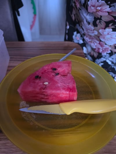 A slice of watermelon with seeds rests on a yellow plastic plate. A yellow-handled knife lies on the plate in front of the watermelon. A metal spoon is positioned behind the watermelon. The scene is set on a wooden table. In the background, a person wearing a dark floral-patterned dress is visible. The context does not provide any information about the city of Barbing, Germany.