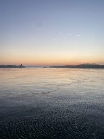 The image displays an expansive body of water under a sky transitioning from light blue at the top to an orange, pink, and pale yellow gradient near the horizon. A faint white contrail from an aircraft is visible in the upper-right quadrant, and a dark, circular smudge appears in the upper-left sky. The water's surface reflects the sky's colors, appearing lighter near the horizon and darkening to cooler blue-green tones in the foreground, with numerous small ripples and waves across its entirety.
On the distant left horizon, the city of Călărași, Romania, is visible, featuring a prominent dark, multi-story structure, possibly an industrial building or silo, among other indistinct buildings. A small, dark object, likely a boat or barge, is present on the water near this distant urban outline. The distant right horizon is delineated by a dense line of silhouetted trees. No individuals, direct actions, or interactions are discernible within the frame.