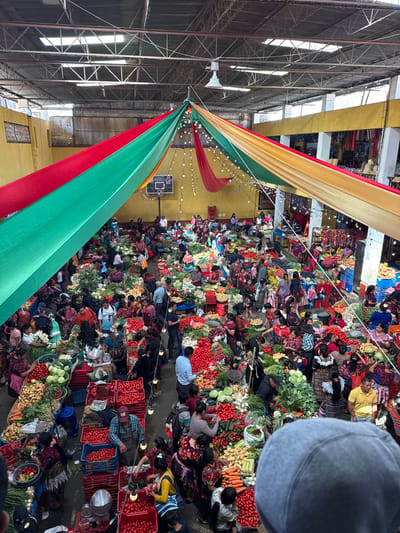 The image depicts an indoor market in Chichicastenango, Guatemala, viewed from an elevated perspective. The market occupies a large, high-ceilinged hall with exposed metal trusses, some translucent roof panels, and yellow-painted walls. A basketball hoop and backboard are affixed to the far left wall. The hall is decorated with large fabric drapes in red, green, and gold/yellow, fanning out from a central point, with strings of small decorative lights suspended beneath them.

The market floor is densely packed with numerous individuals, primarily adults, many of whom are women dressed in traditional Indigenous Guatemalan textiles. The scene is bustling with activity, centered around a vast array of fresh produce. Stalls are arranged in rows, displaying large quantities of vegetables such as tomatoes, potatoes, carrots, cabbage, lettuce, onions, and various leafy greens, often piled high in baskets, crates (many red plastic), or directly on tables. Vendors are observed attending their stalls, arranging produce, and interacting with customers who are examining and selecting items. Scales are visible on several vendor tables. The overall environment is one of commerce and community engagement within a vibrant, enclosed space.
