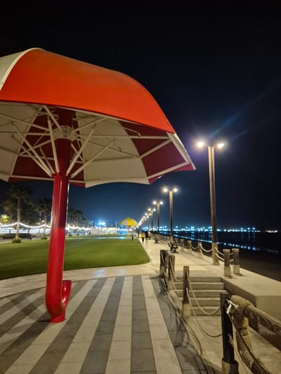 A night scene captures a waterfront promenade in Fujairah, United Arab Emirates. In the foreground, a large red and white umbrella structure stands prominently, its metal framework visible beneath the canopy. The ground is paved with large, rectangular tiles arranged in a striped pattern of light and dark gray. To the left, a well-maintained green lawn extends towards the background, lined with palm trees and decorative string lights.
Further along the promenade, a row of streetlights illuminates the pathway, casting pools of light. Benches are placed along the walkway, offering places to rest. A low railing made of wooden posts and rope separates the promenade from a body of water, where distant lights reflect on the surface, suggesting a cityscape or harbor. Several people are visible walking along the promenade in the mid-ground and background, some appearing to be couples or small groups. The overall atmosphere is one of a lively, well-lit public space at night.