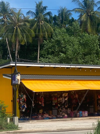 This outdoor shot depicts the exterior of a shop, possibly a souvenir store, located in Baan Nai Suan, Thailand. The shop is characterized by its bright yellow facade and a large, yellow awning. The store is filled with various merchandise, with prices indicated on some items, displayed on shelves. A sign with "Ava Sho" is visible. The scene is set against a backdrop of lush greenery, including palm trees and other trees. The road runs alongside the shop.