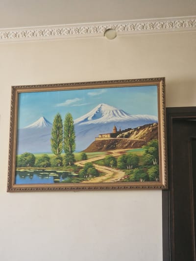 The image shows a framed painting hanging on a wall in Nor Kharberd, Armenia. The painting depicts a landscape with snow-capped mountains in the background, a body of water in the foreground, and trees and a historical building on a hill. The wall is painted a light cream color and has decorative molding along the top. A dark wooden door is visible on the right side of the image.