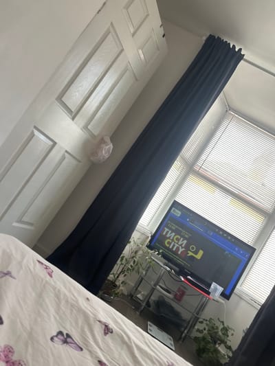 This image depicts an interior room, likely a bedroom or living area, in Boston, United Kingdom, captured from an elevated, angled perspective looking towards a window.

In the immediate foreground, a white duvet with a pattern of purple butterflies is visible, suggesting a bed. To the left, a white paneled interior door is partially open, and a light-colored, crinkled object, resembling a shower cap, hangs from its upper right panel.

Central to the mid-ground, a dark blue curtain hangs from a silver rod, drawn across the upper left portion of a large window. The window itself is fitted with white horizontal blinds, which are partially open, allowing bright daylight to illuminate the room.

Below the window, a black flat-screen television is powered on and resting on a clear glass and metal stand. The television screen displays content in Cyrillic script, featuring "ТИСИ CITY" (TISI CITY), "ОТ ПОНЕДЕЛНИК ДО ПЕТЪК" (MONDAY TO FRIDAY), and times "11:00" and "17:00", along with the URL "www.city.bg". On the top shelf of the stand, next to the TV, a red object and a clear plastic container are visible, along with various cables. On the bottom shelf, a dark-colored device is present, also surrounded by cables.

On the floor to the right of the TV stand, two potted plants with green foliage are visible. To the left of the TV stand and partially obscured by the bed, a white digital weighing scale rests on a medium-toned hard floor surface. The walls of the room are white. No people are present in the scene.