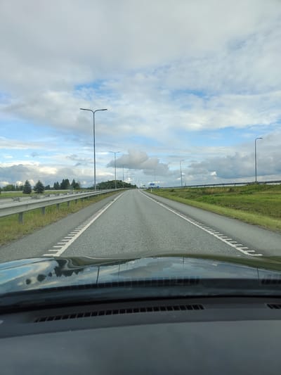 The image depicts a view from inside a vehicle traveling on a multi-lane highway. The highway stretches into the distance, flanked by grassy shoulders and a metal guardrail on the left. Several tall streetlights line the side of the road. The sky is partly cloudy with patches of blue. In the background, a low, tree-covered hill is visible on the right. A sign with blue text is visible on the left side of the highway in the distance. The dashboard of the car is in the foreground, reflecting the sky and trees. The scene appears to be in a rural or semi-rural area outside of a city. Based on the road markings and landscape, it is likely a highway in Estonia. The city of Kukruse, Estonia is a small settlement known for its industrial heritage and shale oil production, and while this image does not directly show any specific landmarks of Kukruse, it is plausible this highway is in its vicinity or leads to it.