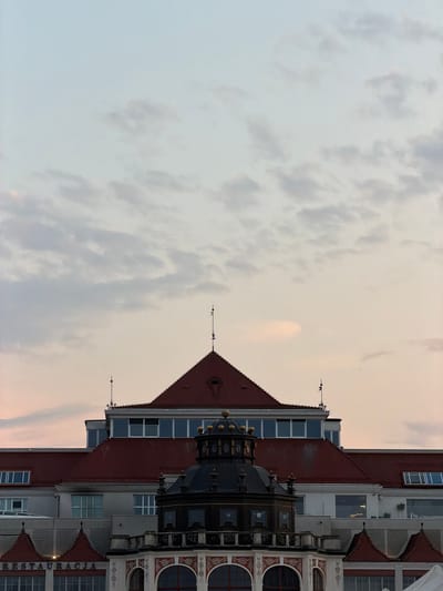 The image shows a close-up view of the historic Grand Hotel in Sopot, Poland, under a cloudy twilight sky. The building features a prominent red-tiled roof, a dark ornate dome, and numerous windows. The architecture suggests a blend of historical styles, with detailed decorative elements visible on the lower facade. The sky is a pale gradient of blues and pinks, with scattered clouds. The overall impression is of an imposing and elegant structure captured during the transitional light of dusk.