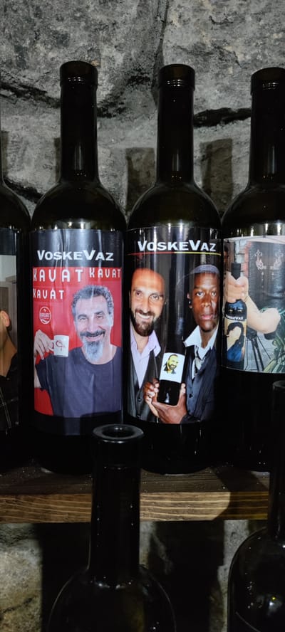 The image shows four dark glass wine bottles lined up on a wooden shelf in front of a textured stone wall. The bottles are labeled with "VoskeVaz" and portraits of people. The first bottle from the left features a man holding a small white cup with a red flamingo logo. The second bottle shows two men, one with a beard and wearing a light-colored shirt, and the other with a darker complexion, wearing a suit. The third bottle displays a person holding a miniature bottle with a portrait on its label. The fourth bottle's label is partially obscured but appears to have a similar style. A fifth bottle, empty and open-mouthed, sits in the foreground on the shelf, and another bottle is visible in the lower right corner. The lighting is dim, creating shadows on the stone wall and highlighting the labels on the bottles.