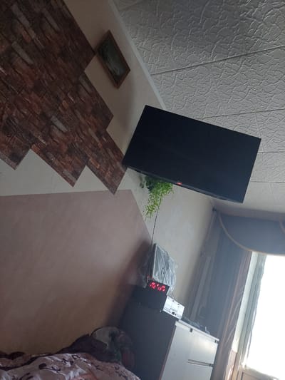This is an indoor scene, likely a bedroom, taken in Cimișlia, Moldova. The room features a textured white ceiling. A large, flat-screen television is mounted on the wall. To the left of the television, a decorative wall panel with a brick pattern is visible. Below the television, on a dark cabinet, sits a digital alarm clock displaying "22 6 25 10 13 0". The room also contains a window with curtains, a dresser with white drawers, and a bed with patterned bedding. A potted plant with long, green leaves hangs from the wall near the television. The overall lighting suggests daytime.