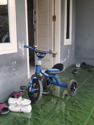 The scene appears to be a partially outdoor area, possibly a porch or patio, in Cibodas, Indonesia, characterized by green tiled flooring with a marbling pattern. Adjacent to the tiles is a gray concrete or painted surface.

In the foreground sits a blue child's tricycle.  Beside the tricycle are several pairs of shoes: white ballet flats and purple sandals. In the background, a line of sandals and flip-flops of various colors are aligned along the green tiled floor. A door with white panels and a rectangular transom window is visible in the middle ground.