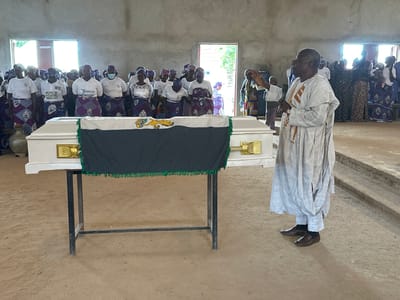 In Kufana, Nigeria, a somber gathering unfolds inside a simple, unpainted building. A white coffin, draped with a dark cloth trimmed in green fringe and featuring a green and yellow emblem, rests on a black metal stand in the foreground. A man in a light-colored traditional robe and pants, his hands raised in a gesture of prayer or blessing, stands to the right of the coffin, facing a congregation of people.

The congregation, primarily women dressed in white tops and vibrant purple and grey patterned skirts and headscarves, stands behind the coffin. Some individuals wear face masks. The background reveals open doorways and windows, hinting at the time of day. The overall atmosphere appears to be one of solemnity and reverence, likely a funeral or memorial service. The ground is a sandy, earth-toned surface. Visible text includes "WhiteCloud" on one of the woman's shirts.