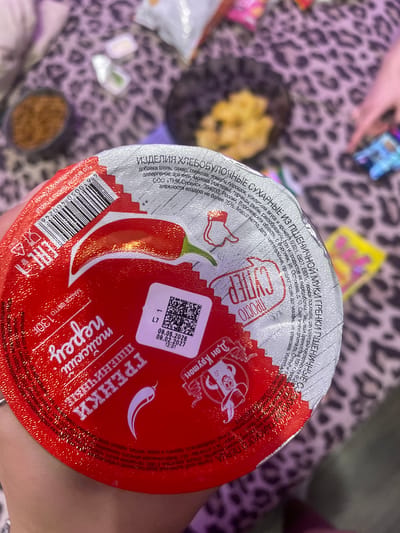 The image shows the lid of a food product with Russian text and a barcode. The lid is red and white with a drawing of a chili pepper. There is also a QR code with text that appears to be a date and time. The background is a blurry leopard print fabric. The text on the lid mentions "ИЗДЕЛИЯ ХЛЕБОБУЛОЧНЫЕ" (bakery products), "МУКА ГРЕНКИ" (flour rusks), and "ПЕРЕЦ" (pepper). The text also includes manufacturing information such as "ООО «ТМК-Сервис»", an address in Voronezh, Russia, and a date of production. There is no visual information in the image that would suggest a location in Zmeinyi, Russia.