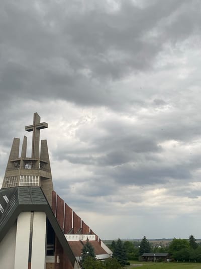 The image shows a modern church with a tall, concrete bell tower topped with a cross. The bell tower has vertical concrete fins and open spaces, with windows on the lower section. The church itself has a distinctive architectural style with a steeply pitched roof covered in dark grey material, white walls, and a section of reddish-brown brickwork. The sky is overcast with heavy, dark grey clouds, suggesting an impending storm. In the background, a rural landscape with trees, fields, and scattered buildings can be seen. The overall impression is one of solemnity and anticipation, due to the dramatic sky and the imposing religious structure. The church is Saint Joseph's Church in Świdnica, Poland. There are no people visible in the image.