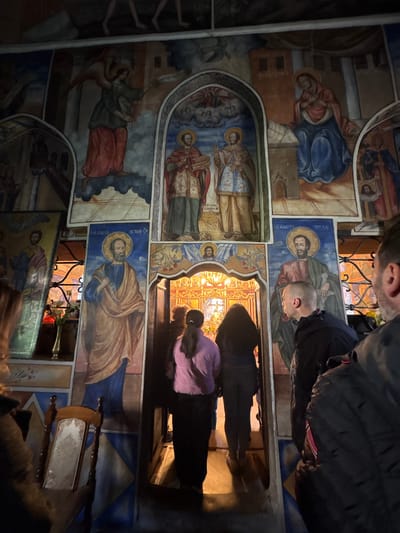 The image depicts the interior of a church featuring a wall covered with religious frescoes, likely in Karadjovo, Bulgaria. The frescoes depict various saints and religious scenes rendered in traditional Byzantine style. Visitors are present, with individuals peering through a doorway or window in the wall. The scene is dimly lit, emphasizing the frescoes' colors and textures. The room appears crowded, with bodies and artifacts partially obscuring the view of the paintings. A lectern stands near the wall.