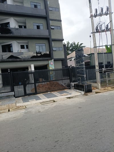A medium shot captures a street view in Rumuokwuta, Nigeria. On the left, a multi-story building with a dark grey facade features staggered balconies and dark-framed windows. The building's exterior walls are a light grey, textured material. In the foreground, a black metal gate runs across the property line. To the right of the building, a tall, black, cylindrical object wrapped in black plastic sheeting and white tape stands in front of a dark fence. Adjacent to this is a utility pole with electrical equipment, including insulators and wires. Behind the fence, houses with brown tiled roofs are visible. The ground in front of the gate is paved with square tiles in a grey and white diamond pattern. The street in the foreground is asphalt, showing some wear and patches of discoloration. The sky is overcast with grey clouds.