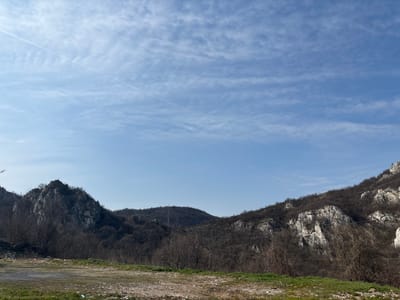 The scene depicts a mountainous landscape in Lyutibrod, Bulgaria, under a clear, light blue sky. The sky, occupying the upper two-thirds of the frame, displays scattered cirrus clouds and a faint, linear contrail extending diagonally from the upper left to the upper right. The foreground comprises uneven ground with patches of dry grass and exposed soil, including a single piece of white debris near the center-bottom. The mid-ground and background are dominated by mountain ranges. A dark, rugged, rocky peak with sparse, bare deciduous trees is prominent on the left, featuring a small, dark, cross-like structure on its summit. To the right, an extensive mountain slope exhibits prominent white rock formations interspersed with dark, leafless trees. Several dark utility lines are visible, traversing the mid-section of the right mountain's slope. No human or animal subjects are present, and no actions or interactions are discernible. The vegetation indicates a dormant season.