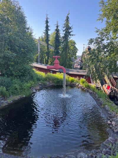 The image captures a scene at Särkänniemi amusement park in Tampere, Finland, featuring a water ride with a prominent red faucet sculpture that acts as a fountain. In the foreground, a dark, rippling body of water is illuminated by the sunlight, with a stream of water cascading from the faucet and creating splashes. The faucet sculpture, a large, bright red object, is positioned to the left of the frame, with its spout directed downwards towards the water. Behind the faucet, a red-roofed building is visible, surrounded by lush green foliage and trees. In the background, the metallic structure of a roller coaster stretches across the sky, its tracks winding through the trees. To the right of the frame, a boat carrying several passengers is descending a track, indicating the presence of an active amusement park ride. The sky is clear and blue, suggesting a sunny day. The overall setting is an outdoor amusement park environment with natural elements like trees and water integrated with man-made attractions.