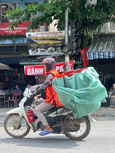 A man in an orange vest and grey hooded sweatshirt rides a white scooter with a large, light green tarp secured to the back. The tarp appears to be filled with items. He is wearing light grey pants and blue open-toed shoes. A red cylindrical container is attached to the side of the scooter. The scooter is on a paved road with a yellow line. In the background, there are buildings, signs, trees, and a traffic light. One sign, in red and white, reads "THƯƠNG PHƯƠNG". Another sign, partially visible, features an image of a landscape and Vietnamese text. A partial blue and white striped awning is visible over a building entrance. The scene appears to be in a commercial area in Tân Quang Commune, Vietnam, indicated by the signage and urban setting.