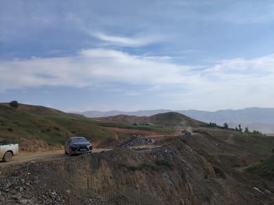 The image depicts a hilly, rural landscape in Farab, Tajikistan, under a partly cloudy sky, suggesting daytime. The scene is characterized by rolling green hills interspersed with areas of disturbed earth, indicating construction or excavation activities.
Several vehicles are present, including a dark-colored SUV parked on a dirt road and a white pickup truck to its left. In the distance, construction equipment, including a bulldozer and a truck, can be seen working on a road or clearing land, with dust rising from their operations. A blue container is also visible near the construction site.
The terrain is uneven, with steep inclines and rocky patches, typical of a mountainous or arid region. The vegetation is sparse in some areas, with grass covering the greener hillsides. The distant mountains are hazy, suggesting a considerable distance and possibly atmospheric conditions. The overall impression is one of ongoing development or resource extraction in a natural, undeveloped setting. No people are clearly visible. The license plate of the SUV reads "5882 AKB".