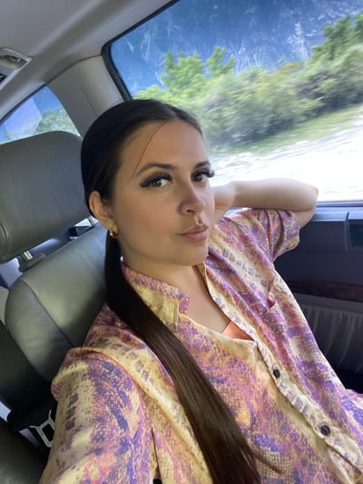 A young woman with dark hair, long eyelashes, and a gold hoop earring is seated in the passenger seat of a vehicle. She is wearing a patterned short-sleeved shirt with shades of purple, yellow, and pink, and a coral-colored tank top underneath. Her left arm is resting on the back of her head, and her right hand is near the window. The interior of the car, including a gray leather seat and part of the dashboard, is visible. Through the car windows, a blurred landscape of green trees and mountains under a blue sky can be seen, suggesting the vehicle is in motion through a scenic, possibly mountainous, area outside the city of Irganay, Russia.