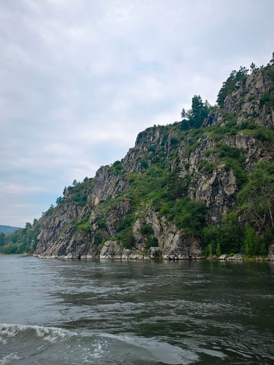 The image depicts a wide, dark river flowing in the foreground. On the right side of the image, a steep, rocky mountain rises dramatically from the riverbank, covered in lush green vegetation and scattered pine trees. The mountain's surface is rugged and uneven, with visible rock formations and crevices. The sky above is overcast with soft, grey clouds, creating a muted lighting effect. The left side of the image shows a more distant, tree-covered shoreline, suggesting a natural, undeveloped landscape. The overall scene conveys a sense of natural grandeur and remoteness. The location is the Yenisei River near the village of Atamanovo, Krasnoyarsk Krai, Russia.