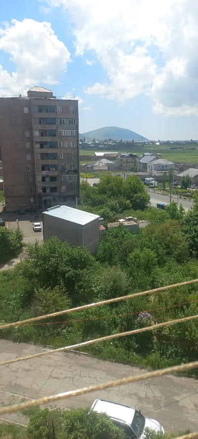 The image captures a view of a residential area in the city of Sevan, Armenia, on a bright, partly cloudy day. In the foreground, a cracked asphalt surface with some grass is visible, with prominent yellow and red wires strung across it. A white van is parked on the asphalt, partially obscured by green foliage.
Dominating the left side of the midground is a tall, multi-story apartment building constructed of concrete and reddish-brown stone. The building has numerous windows and balconies, some of which are adorned with laundry hanging out to dry. In front of the apartment building, a smaller, rectangular structure with a pitched, metallic roof stands. A white car is parked to the left of this structure, and another car is visible further back near the building.
The background reveals a suburban landscape with numerous houses, many featuring grey roofs, interspersed with green trees and fields. A paved road winds through the area, with a few vehicles visible. In the far distance, a large, rounded mountain forms the horizon under a blue sky filled with scattered white clouds. The overall impression is one of a typical day in a residential neighborhood in Sevan.