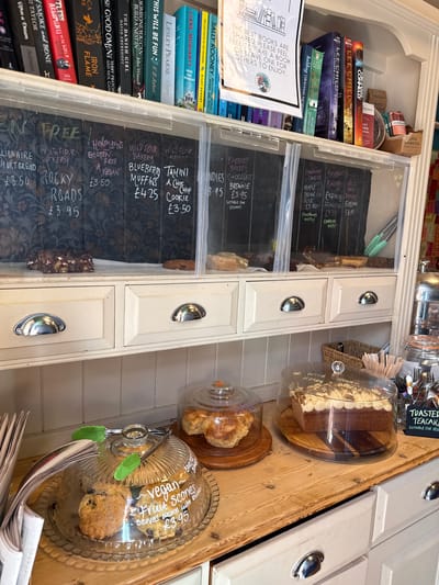 This image depicts a cozy bookshop and cafe located in West Oxfordshire, United Kingdom. A selection of pre-owned books, including works by Lee Child, Sally Rooney, and Neil Gaiman, is displayed on the left side of the image. Adjacent to the books is a wooden shelving unit with drawers containing various baked goods behind transparent covers, such as brownies, muffins, and flapjacks, with chalkboard labels indicating their ingredients and prices. Cakes and scones, some labeled "vegan," are arranged on wooden platters under glass domes on a wooden counter, next to a stack of newspapers. Serving utensils are attached to the scones' display, and a sign promotes "Toasted Teacake." The overall impression is one of a charming, independent establishment with a focus on literature and homemade treats.
