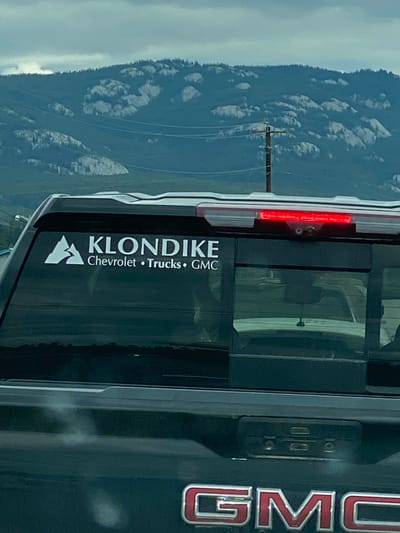 The image captures a rear view of a dark-colored GMC pickup truck, parked in what appears to be an outdoor setting with a mountainous, cloudy backdrop. The truck has a decal on its rear window that reads "KLONDIKE Chevrolet • Trucks • GMC" with a white mountain logo. The truck's third brake light is illuminated red. In the background, a wooden utility pole with wires is visible against a backdrop of rugged, rocky hills and a muted sky. The overall scene suggests a location within or near a natural landscape, possibly on the outskirts of Whitehorse, Canada, given the "Klondike" branding which is strongly associated with the Yukon Territory. There are no people visible in the image.
