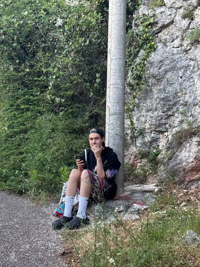 A young person is sitting on a rock next to a concrete pole, holding a phone. They are wearing a black hoodie with purple floral accents on the sleeves, colorful shorts, white socks, and black platform shoes. Their hair is short and they have a black baseball cap on. The background consists of lush green foliage and a rocky cliff face. The ground is covered in gravel and grass. The scene appears to be outdoors, possibly in a natural or park setting.