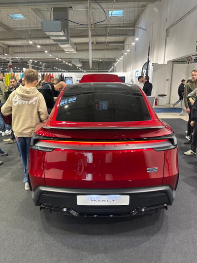 The scene depicts the rear view of a deep red Tesla Model Y electric vehicle positioned on a dark grey carpeted floor within a brightly lit indoor exhibition hall in Wolica, Poland. The vehicle's continuous horizontal rear LED light strip is illuminated. Below it, the word "TESLA" is embossed in chrome lettering. A small emblem resembling the Union Jack flag is affixed to the lower right side of the tailgate. A white temporary license plate displaying "MODEL Y" is mounted on the lower black bumper. The rear window is dark-tinted, reflecting overhead lights and some interior details.

Numerous individuals are present around the vehicle. In the immediate foreground, a young male wearing a beige hooded sweatshirt and blue jeans stands facing the car, his back to the viewer. To the left, several adult males are gathered, some leaning forward, appearing to observe the car's interior. In the background, more people are visible, some walking, others standing by various exhibition displays. The spacious hall features a high white ceiling with exposed silver ventilation ducts and multiple overhead lighting fixtures. Rectangular skylights are also visible in the ceiling. Background elements include displays of what appear to be material samples on the left, a partially obscured red vehicle with an open hood, and white walls with emergency signage further to the right.