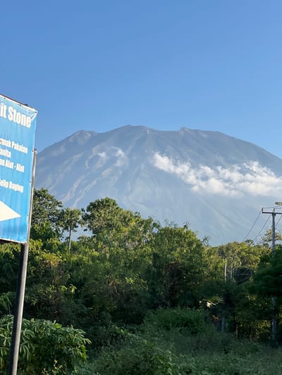 This is a photograph of Mount Agung, an active volcano located in the Karangasem regency in eastern Bali, Indonesia, as seen from Kubu, a village on Bali's northeast coast. The image features the majestic mountain dominating the background, with some clouds clinging to its slopes. In the foreground, there is lush green vegetation, including trees and shrubs, suggesting a tropical environment. To the left, a blue signpost with white text is partially visible, indicating a street-level view and possibly a local business or landmark. The sky above is clear and blue, typical of a sunny day. The overall scene conveys a sense of natural beauty and tranquility, with the imposing volcano serving as a striking focal point.