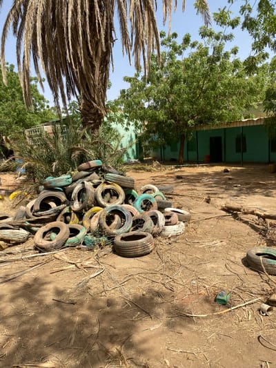 The image shows a pile of discarded tires in front of a building in Khartoum, Sudan, located at latitude 15.552 and longitude 32.57. The building is light blue with white trim and has a door and several windows. There is a large palm tree in the foreground and a smaller tree to the left of the building. The ground is covered in dirt and dry grass. There are no people present. The image appears to have been taken on a sunny day.