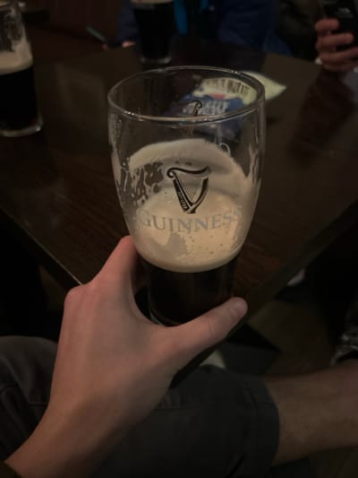 A partially consumed pint of Guinness sits on a dark, likely wooden, surface. The glass, adorned with the Guinness logo, is held by a person's hand. The stout's creamy head contrasts with the dark liquid. Another glass, similarly filled, is visible nearby, suggesting the setting is a pub or social gathering. The overall composition is slightly tilted, adding a casual, perhaps even celebratory, feel.