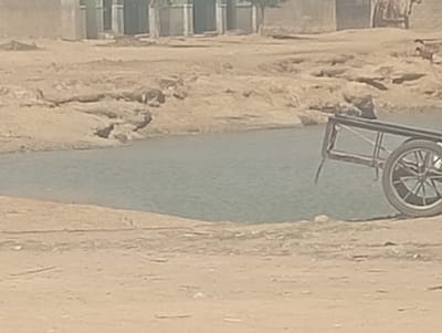 The image displays an outdoor scene in Sumaila, Nigeria, characterized by a dry, arid environment under daylight. A calm body of water, appearing light blue-grey, occupies the central portion of the frame, flanked by irregular, light brown earthen banks. On the right bank, a two-wheeled cart with a dark frame and a single visible spoked wheel is positioned, its rear oriented towards the viewer and angled towards the water. A dark object, possibly a container or bag, rests on the cart's platform or immediately adjacent to it. The immediate foreground consists of flat, dusty, light brown earth. In the distant background, beyond the water and an elevated, eroded earthen area, multiple light-colored structures, resembling incomplete buildings with exposed concrete or blockwork, are visible. No individuals are present.