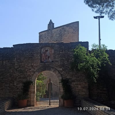 The image shows a stone archway in the city of Terlizzi, Italy. The archway is part of a stone wall, and above it, a small stone structure resembling a bell tower is visible against a clear blue sky. A colorful fresco is embedded in the stone wall above the archway, depicting figures in religious attire. To the right of the archway, a lush green tree grows against the wall, and a tall lamppost stands nearby. In front of the archway, a black metal gate is visible, with sunlight casting shadows on the ground. Two terracotta pots with plants are placed to the left and right of the archway. The bottom right corner of the image displays a date and time stamp: "10.07.2026 08:34". The overall scene is an outdoor environment, likely a historical site or a church, characterized by ancient stone architecture and vegetation.