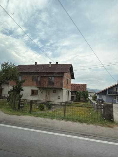 The image depicts a street scene in Putićevo, Bosnia and Herzegovina, on a cloudy day. A two-story house with a terracotta tile roof and exposed brick on the upper level is prominently featured. The house has a balcony with a metal railing on the second floor. Adjacent to it, a smaller building with a similar roof is visible. Further down the street, another house with a grey facade and a darker roof is seen, with a dark-colored car parked in front of it. A metal fence runs along the front of the properties, separating them from the road. The road itself has a white line marking. Green trees and vegetation are scattered throughout the scene, and a field with yellow flowers is visible behind the fence. The sky is overcast with scattered clouds.