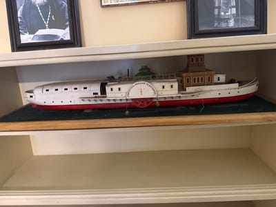 The image shows a model of a steamboat on a shelf. The steamboat is white with a red hull and a wooden superstructure. It has a paddle wheel at the stern and a green dome on top. There is text written on the paddle wheel in Cyrillic letters that appears to read "СВЯТИТЕЛЬ НИКОЛАЙ ЧУДОТВОРЕЦ" which translates to "Saint Nicholas the Wonderworker". Above the steamboat model, on another shelf, are two framed pictures. The picture on the left is a black and white photograph of a man in religious attire, possibly a bishop, seated at a desk with a book. The picture on the right is also a framed image, but it is partially obscured and appears to be a black and white photograph as well, possibly of a person in formal or religious garb. The shelves are light-colored, and the wall behind them is a pale yellow. The overall scene appears to be a display of historical or religious artifacts. This scene is likely located in Moscow, Russia, given the Cyrillic text and the religious iconography.