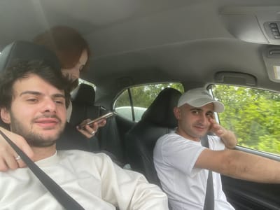 Two men are sitting in the front seats of a car. The man on the left is wearing a white t-shirt and a white baseball cap, resting his head on his hand. The man on the right is wearing a white sweater and a seatbelt, holding a phone. The background shows a blurry view of green trees outside the car window. The interior of the car is visible, including the seats and ceiling. There is no explicit evidence to confirm the city or location of Changilari, Georgia.