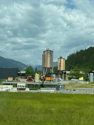 The image depicts an industrial area in Flachauwinkl, Austria, with two tall cylindrical silos dominating the center. The silos appear to be constructed from wood or a similar material, with a metallic lattice framework supporting them. One silo is slightly taller than the other, and both have conical tops with visible piping.
In the foreground, a lush green meadow stretches across the bottom of the frame, dotted with small wildflowers. Beyond the meadow, a construction site is visible, featuring concrete barriers, industrial machinery including what appears to be a truck with a yellow and red warning sign, and several shipping containers. A pale yellow building with a dark roof is situated behind the machinery, and a larger, dark gray building stands to the left.
In the background, rolling green hills covered in dense evergreen trees rise up, with the majestic, layered peaks of the Austrian Alps visible in the distance under a cloudy sky. The clouds are a mix of bright white cumulus and darker, more diffused formations, suggesting an overcast or partly cloudy day. A white car is parked in front of the yellow building. The overall scene suggests an active industrial or construction zone in a mountainous, rural setting.