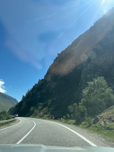 The image depicts a winding paved road through a mountainous region, likely in or near Upper Baksan, Russia. The road curves to the left, with a metal guardrail on the left side. A white van is visible driving away on the road in the distance. The right side of the road is a steep, rocky mountainside covered with dark green coniferous trees and some deciduous trees. The sun is shining brightly from the upper right corner of the frame, creating lens flares and casting light and shadows on the mountain. The sky is clear and blue. The foreground shows the top of a vehicle's dashboard. The overall scene suggests a scenic drive in a rugged, natural environment.
