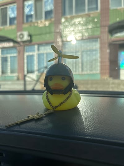 The image shows a yellow rubber duck with a black helmet and propeller on its head, and a gold chain around its neck. To the left of the duck, a toy rifle is placed on a dark, textured surface. The duck and rifle appear to be on the dashboard of a vehicle, with a blurred background of a building and windows, suggesting an urban setting. The overall lighting indicates it is daytime. There are no people present in the image. The location is identified as Konopliane, Ukraine.