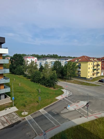 The image shows a residential area in České Budějovice, Czechia. In the foreground, there is a street intersection with a pedestrian crossing and traffic islands. Several apartment buildings are visible in the background, along with trees and a grassy lawn. The sky is blue with some clouds.