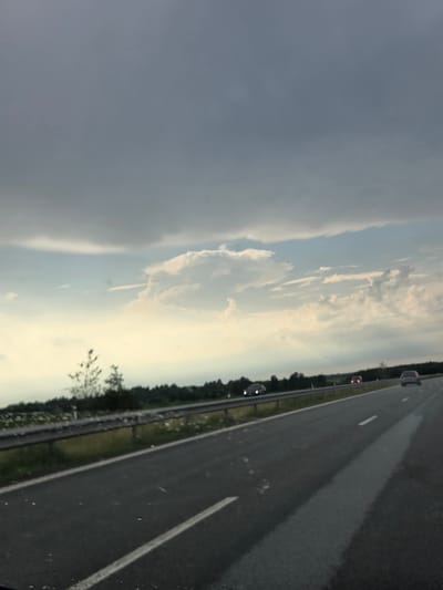 The image depicts a view from a vehicle traveling on a highway in Gudulinė, Lithuania. The scene is dominated by a vast sky filled with dramatic clouds, ranging from dark, ominous gray on the left to lighter, scattered white clouds illuminated by a soft, diffused light on the right. The horizon is visible as a thin line of trees separating the sky from the road.
The activity taking place is travel, indicated by the perspective from a moving vehicle. The road itself is asphalt, with clear white lane markings. Several vehicles are visible on the highway: a dark-colored car in the distance on the right lane, a red vehicle with its brake lights on in the middle distance on the left lane, and another dark vehicle further back on the left lane. A metal guardrail runs alongside the road, separating it from a grassy verge and the trees beyond. Small trees and foliage are visible along the side of the road.
The time of day appears to be late afternoon or early evening, given the soft, low-angled light and the mix of dark and bright clouds, suggesting changing weather conditions. The weather seems to be transitioning, with the darker clouds hinting at potential rain, contrasting with the brighter, clearer sky on the right. There are no visible emotional cues or text in the image.