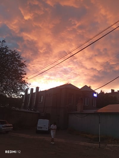 This is a photograph of a dramatic sunset over a town, likely Vedi, Armenia. The sky is filled with vibrant orange and pink clouds, indicating the sun is low on the horizon. Silhouetted against the colorful sky are power lines and the rooftops of buildings. One building in the mid-ground has a glowing blue sign with a white outline of a stylized "L." A white van is parked in front of this building, and a person wearing a white shirt and shorts stands nearby. Another vehicle is partially visible in the lower right corner. The overall mood of the image is serene and beautiful, capturing the tranquil end of a day. The text "REDMI 12" is visible in the bottom right corner, indicating the device used to take the photo.