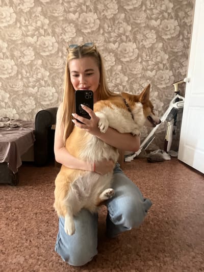 A person is taking a selfie with their Corgi dog. The person is blonde and has sunglasses on their head. The dog is a fawn and white Corgi. They are sitting on a cork floor in a room with floral wallpaper. A white stick vacuum cleaner and a black armchair are visible in the background. The overall mood appears to be relaxed and affectionate. The photo is taken indoors, suggesting it is daytime. There is no visible text in the image.