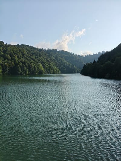 The image depicts a serene lake surrounded by lush green hills and dense forests. The water surface is rippled, suggesting a gentle breeze. The sky is clear with a few wispy clouds. The scene appears to be a natural landscape, possibly a national park or a remote wilderness area. The location is identified as the city of Kharistvala, Georgia, known for its natural beauty and mountainous terrain. The image captures the tranquil atmosphere of this picturesque setting, inviting viewers to appreciate the untouched splendor of nature.
