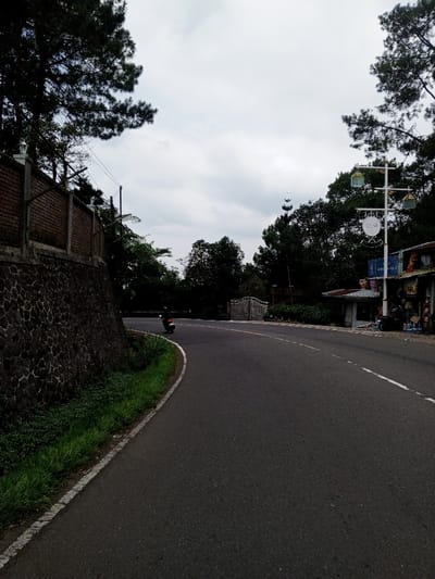 The scene depicts a curved paved road in Ciloto, Indonesia, under an overcast sky. The dark asphalt road curves sharply to the right, marked by a solid white line along its left edge and a broken white line alongside a solid yellow line on its right.
To the left of the road, a dark stone retaining wall supports an elevated area, topped by a reddish-brown brick wall. Above the brick wall, dark green pine trees extend into the sky, and white ornate lamp posts are visible along the wall. Utility poles and wires are also present. Green grass and foliage line the road's edge beside the retaining wall.
To the right of the road, a series of small commercial structures or stalls with various signs are visible. A prominent white multi-arm lamp post with three green and gold lantern-style lights and a circular white sign stands in front of these buildings. Dense dark green trees form a backdrop behind these structures. Further down the road, past the curve, a light-colored decorative gate or archway is visible. A tall, slender spire or antenna structure rises above the distant trees against the cloudy sky.
One red scooter or motorcycle is captured in motion on the left side of the road, proceeding away from the viewer. No individuals are explicitly visible in the scene.