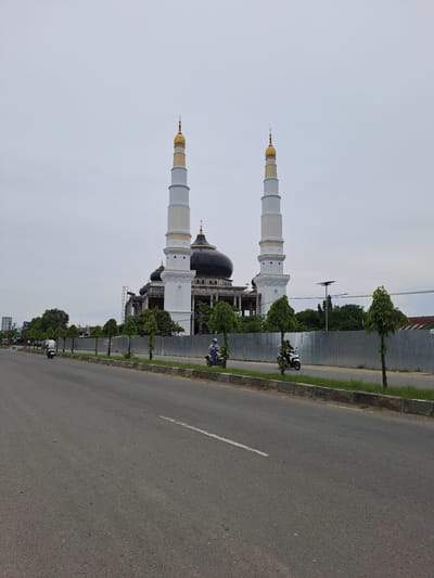 The image shows a mosque with two tall, white minarets topped with gold domes, and a large black dome in the center. The mosque appears to be under construction or renovation, with scaffolding visible behind it. In front of the mosque is a road with a few motorcycles on it, and a line of small trees. The sky is overcast. The location is identified as Pantegajah, Indonesia.
