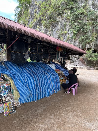 A person is sitting on a pink plastic chair in front of a market stall that is covered with blue and white striped tarpaulin. The stall is located in front of a large limestone cliff with vegetation growing on it. The market stall appears to be selling souvenirs, with items such as postcards and keychains visible. The ground is covered in gravel. The person is wearing black clothing and is holding a phone. The location is identified as a market stall in Krasom, Thailand.