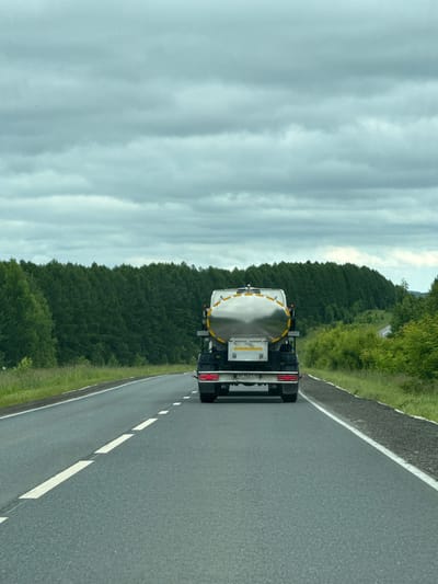 An eye-level, medium shot captures the rear of a silver tanker truck driving on a two-lane asphalt road under a cloudy sky. The truck, with its cargo tank prominently featured, is in the right lane. The road has white dashed lines marking the lanes. On the left side of the road, a grassy shoulder meets a dense forest of evergreen trees. The right side of the road also has a grassy shoulder and a less dense forest with lighter green foliage. In the distance, the road curves to the right, disappearing behind a hill. The scene is set in an outdoor, rural environment, with no people visible. The weather appears overcast. The location is identified as Novaya Kazmaska, Russia.