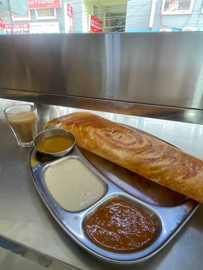 An outdoor shot captures a south Indian breakfast. A metal tray holds a golden-brown masala dosa, its crispy texture evident. Accompanying the dosa are three small metal bowls containing chutneys. One bowl holds a pale, creamy white chutney, another a yellowish sambar, and the third a rich, reddish-brown chutney. To the left, a clear glass tumbler contains a frothy, milk-tea beverage. The food is presented on a stainless steel countertop, with a reflective metal partition behind it. In the background, blurred buildings and electrical wires suggest an urban setting. The scene is set in Kurnool, India.