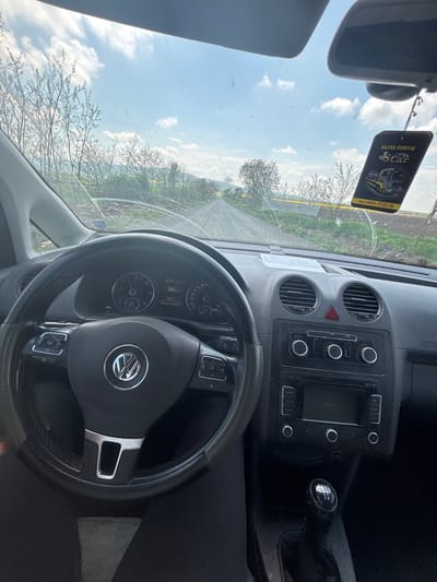 The image shows the interior of a car, specifically the driver's seat. A steering wheel with a Volkswagen logo is in the foreground. The dashboard, air vents, a radio unit, and a gear shift are visible. The speedometer reads 66 km/h. An air freshener hangs from the rearview mirror with the text "ПЪТНА ПОМОЩ JS Car тел: 0890 31 32 30". The outside view through the windshield shows a road, trees, and a partly cloudy sky. Based on the information from the air freshener and the road view, the location can be inferred to be in Podvis, Bulgaria.